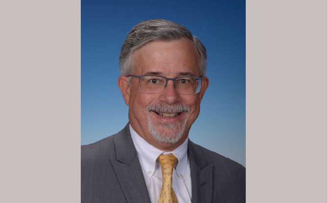 Headshot of man wearing a white collared shirt with gold tie and grey suit jacket in front of blue background