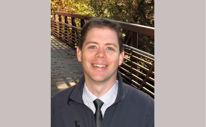 Headshot of man with black tie in front of a bridge