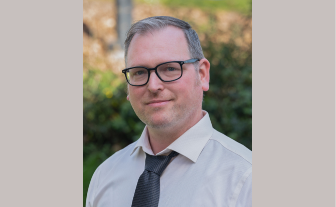 Headshot of a man with white collared shirt and grey tie with glasses standing in front of a natural landscape
