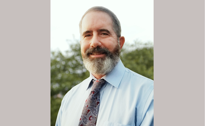 Headshot of man in blue shirt and paisley tie in front of a natural landscape