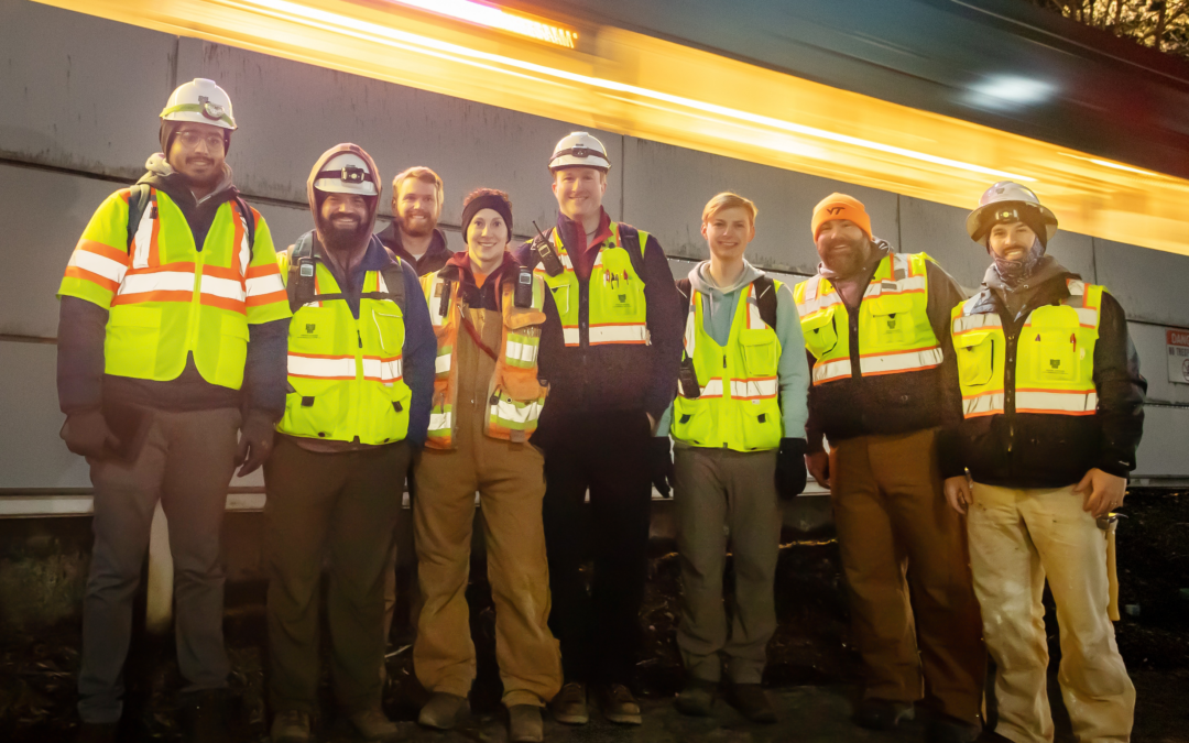 A team of 8 bridge inspectors standing in front of the project site (a tunnel) wearing high visibility vests