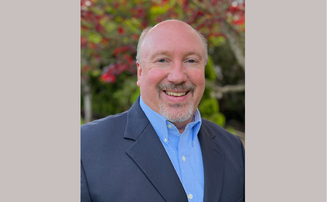 Headshot of a man in a blue shirt with grey suit jacket in front of a flowering tree