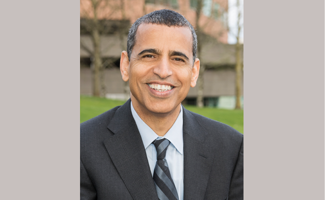 Headshot of man in a suit and tie, with gray hair, in front of a brick building.