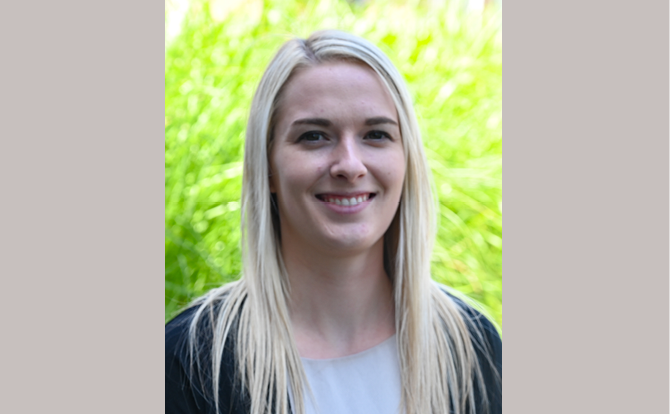 Headshot of a woman with blonde hair, white shirt, and black cardigan in front of a green bush.