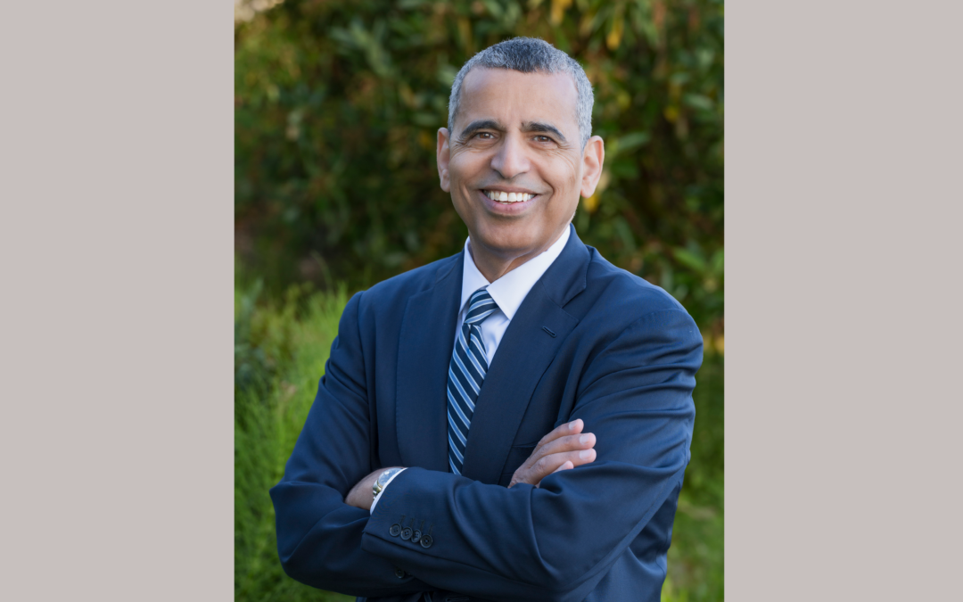 Headshot of a man with a blue blazer and blue striped tie, who is smiling in front of a natural background.