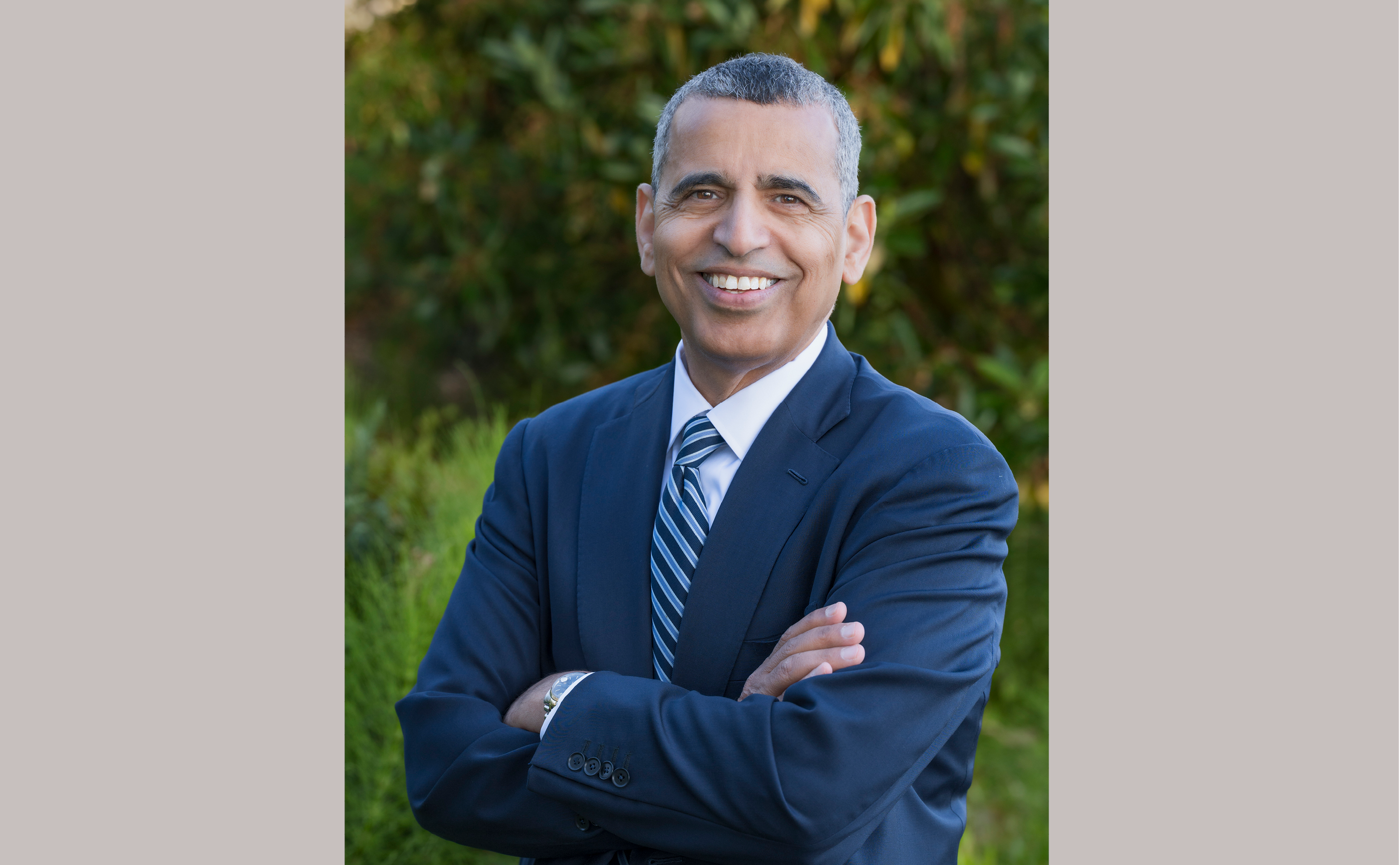 Headshot of a man with a blue blazer and blue striped tie, who is smiling in front of a natural background.