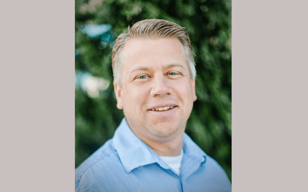 Headshot of a man in a blue button up shirt in front of a natural background.