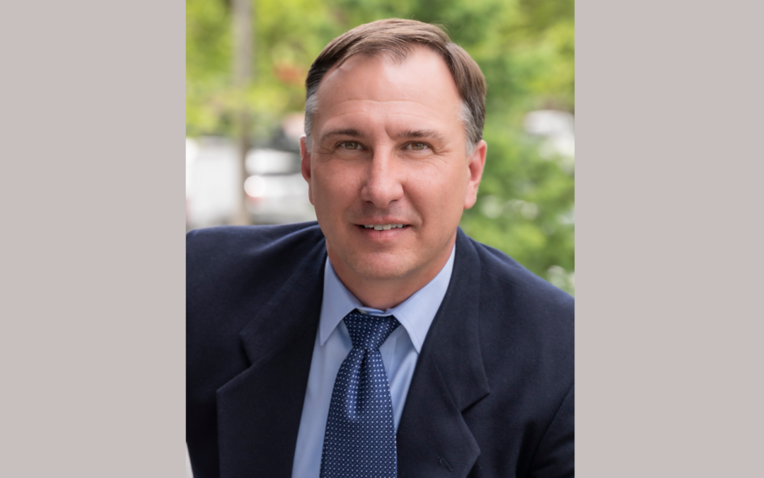 Headshot of a man with a blue shirt, blue tie, and blazer in front of some natural foliage.